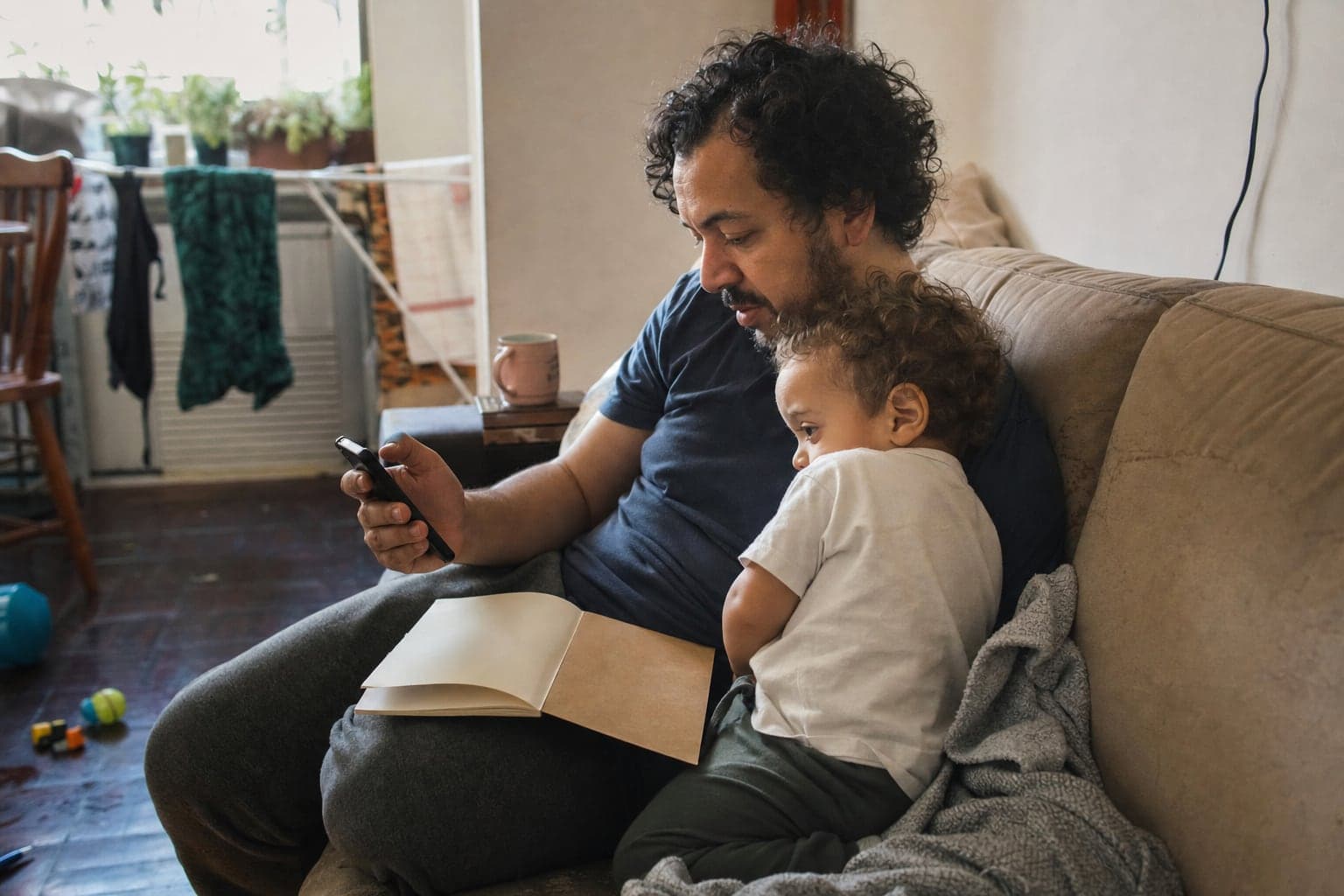 Pai sentado no sofá com o filho pequeno no colo olha o celular, com uma caderneta aberta no joelho
