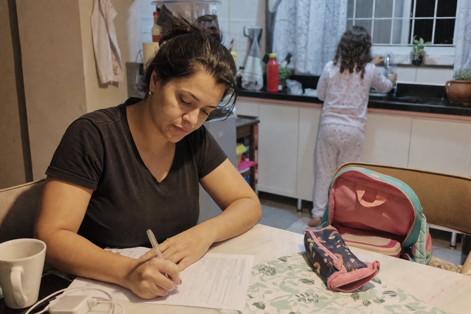 Mãe preenche uma folha à caneta na mesa da cozinha, com a mochila escolar aberta na cadeira ao lado