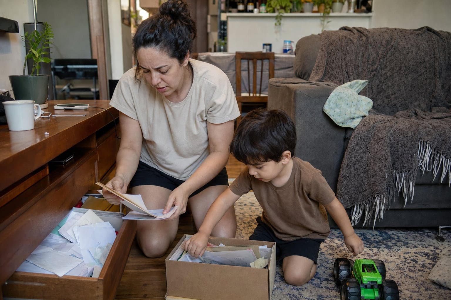 Mãe procura papéis numa gaveta da sala enquanto o filho pequeno mexe numa caixa no chão ao lado